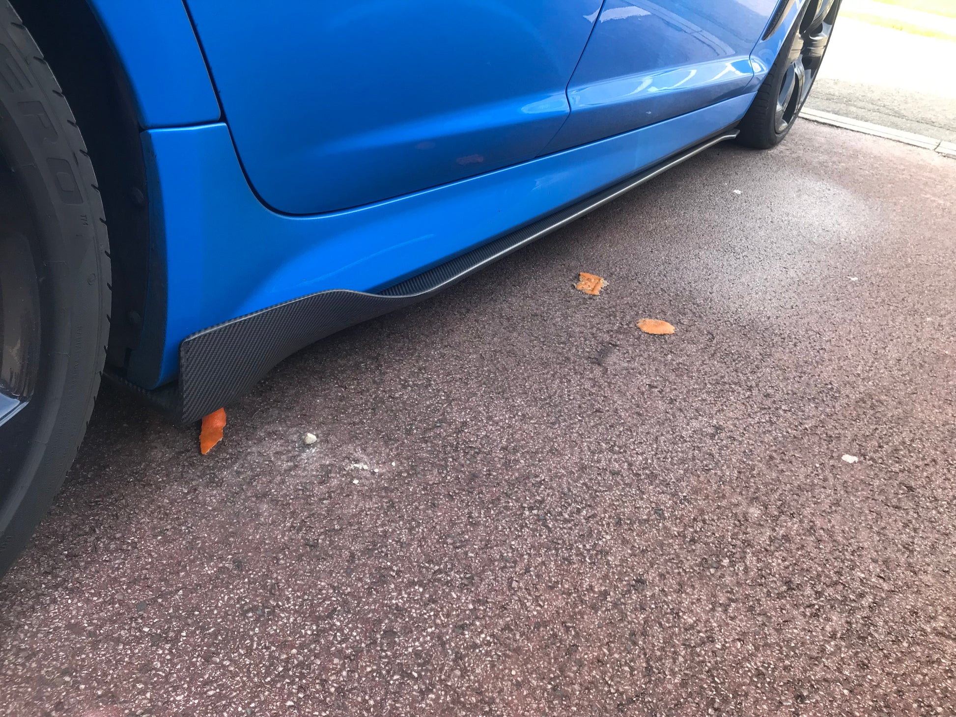 Black carbon fibre side skirts fitted to a blue vehicle, showing the lower section of the car's doors and fenders.