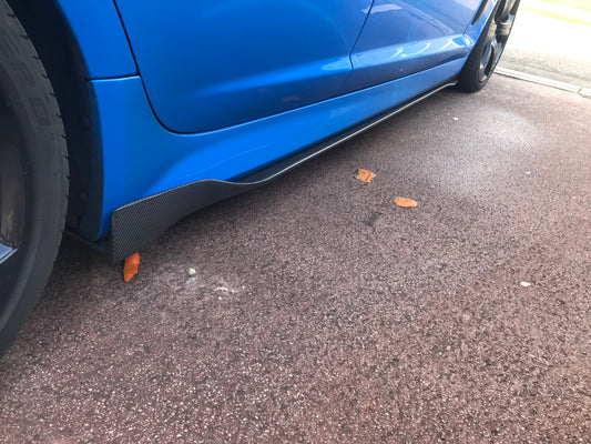Black carbon fibre side skirts fitted to a blue vehicle, showing the lower section of the car's doors and fenders.