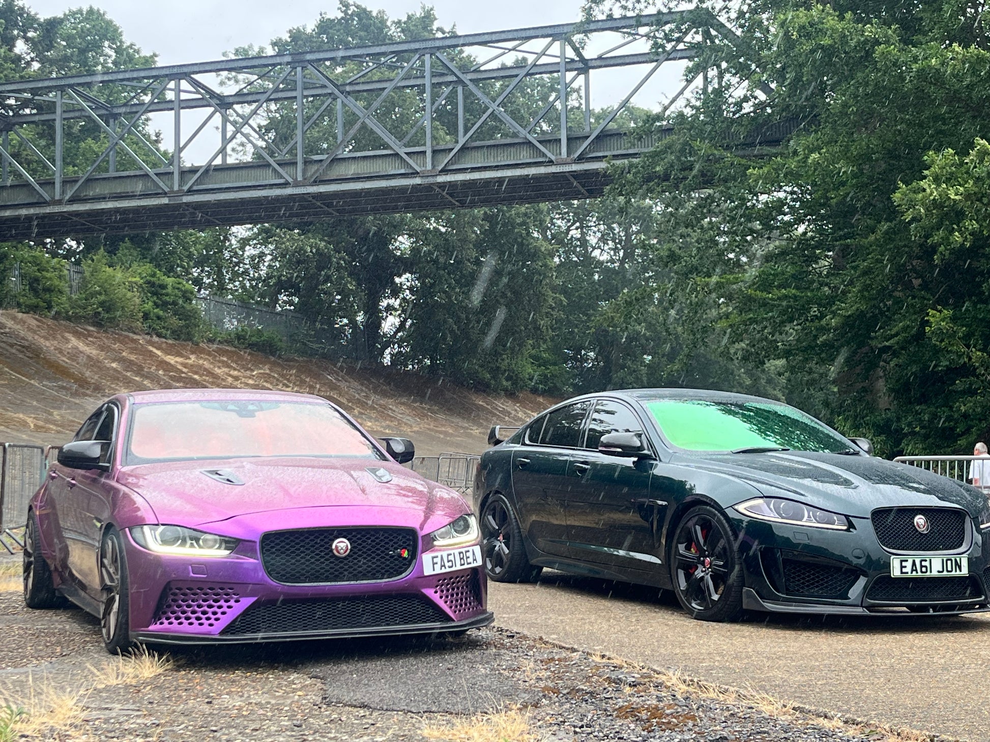 Two Jaguar cars, one purple and one black, parked on a road with a bridge and trees in the background.