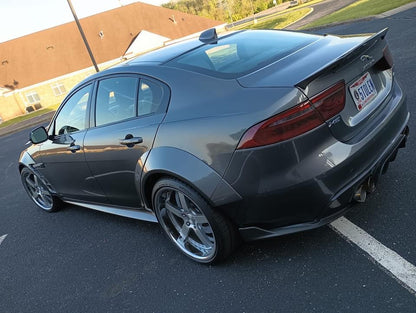 silver jaguar xe with aftermarket carbon fiber rear boot spoiler, car in a parking lot with a brown building in the background